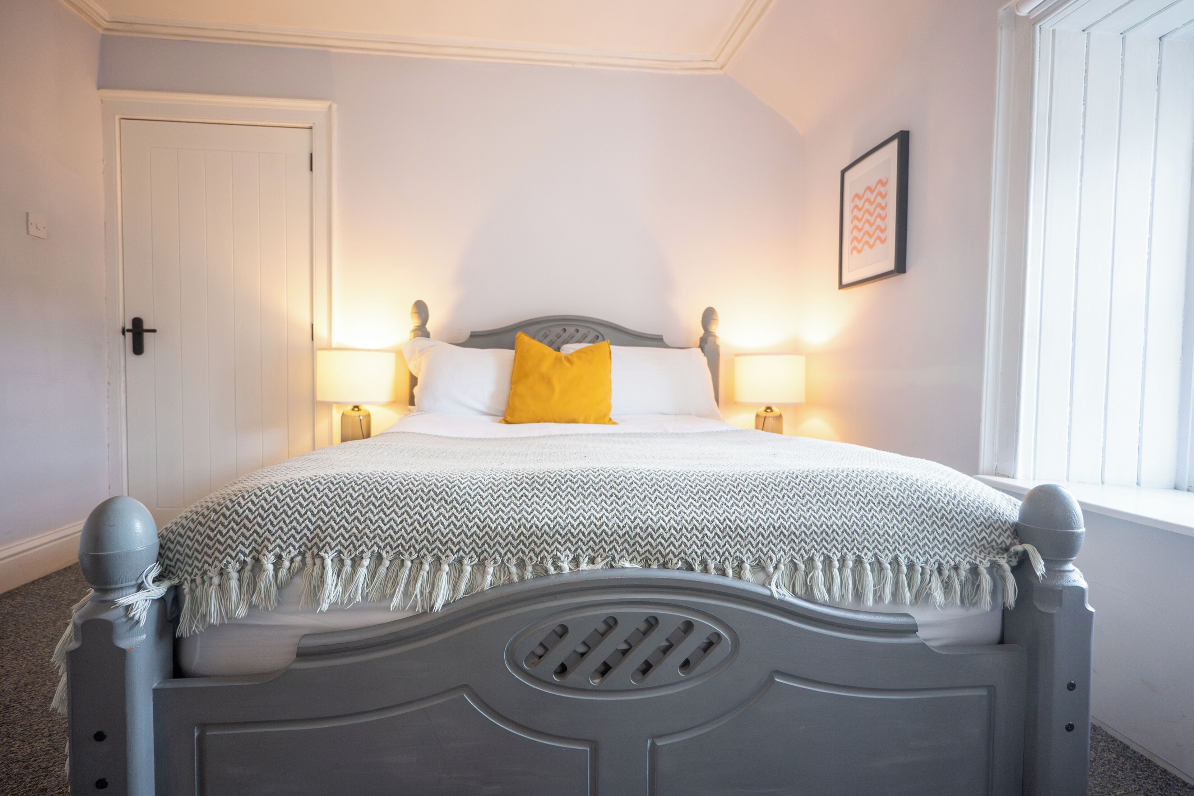 Un letto ben rifatto con una testiera grigia, biancheria da letto bianca e un cuscino giallo decorativo. Sono presenti due lampade ai comodini e un quadro appeso al muro.