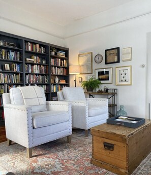 A living room with two white armchairs, a wooden coffee table, a bookshelf filled with books, and a patterned rug.