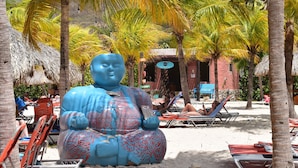 Een groot blauw Boeddhabeeld in een tropische omgeving met palmbomen, strandstoelen en een gebouw op de achtergrond.