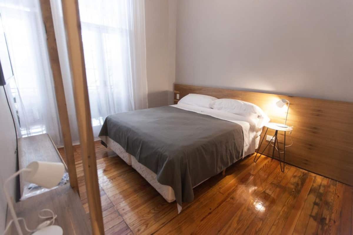 Une chambre à coucher bien rangée, avec une tête de lit en bois, un lit recouvert d’une couverture grise, une lampe de chevet et un plancher en bois.