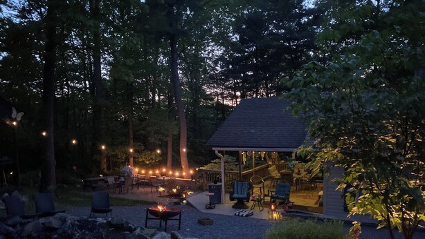 Un'area salotto esterna riparata con sedute in pietra e un braciere, circondata da alberi e luci decorative.