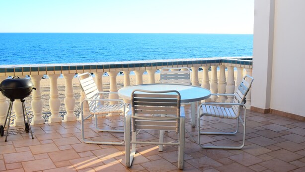 A balcony with a view of the sea, a round table, four chairs, and a grill.