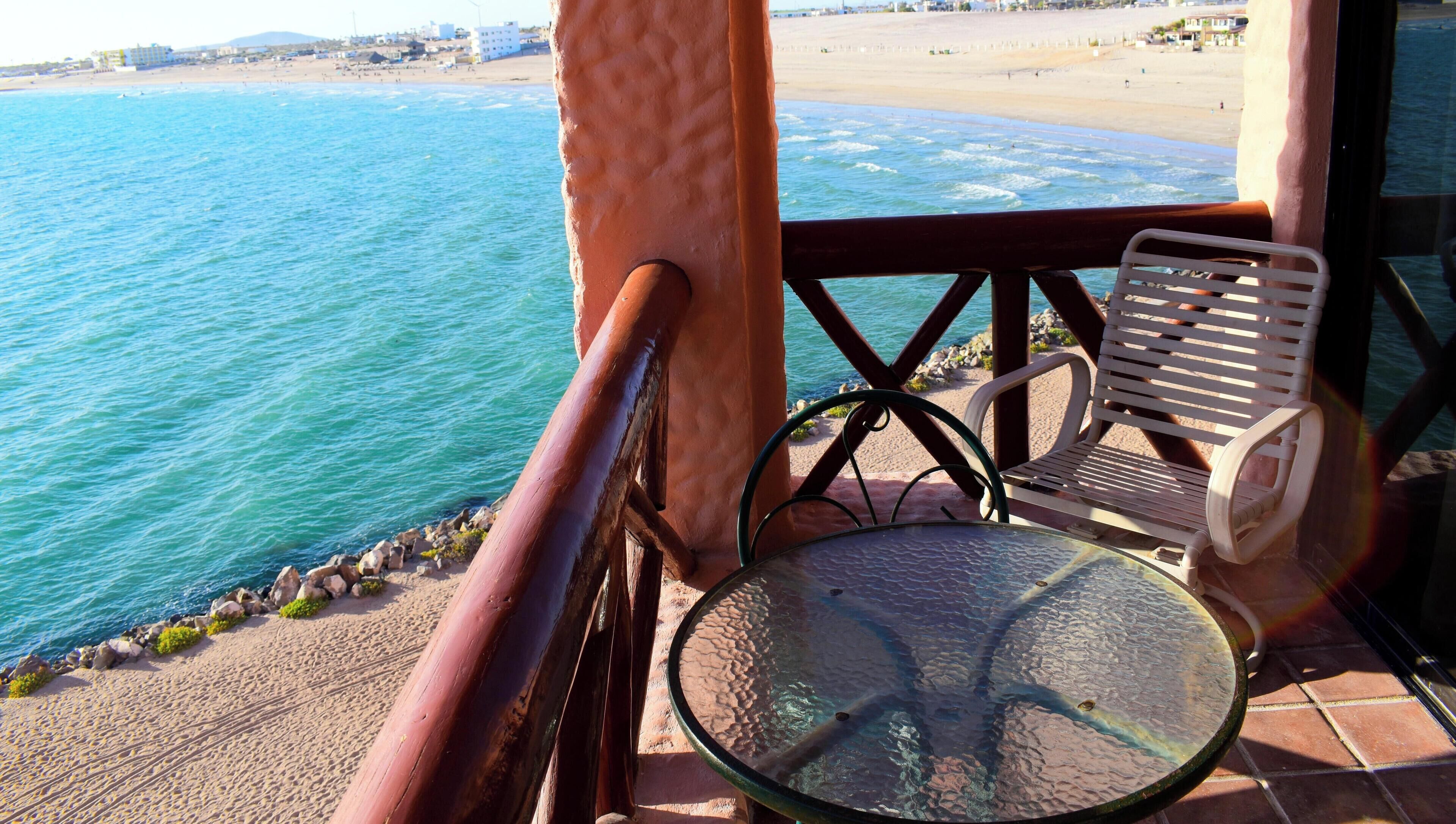 A balcony with a glass table and two chairs overlooking a beach and the sea.