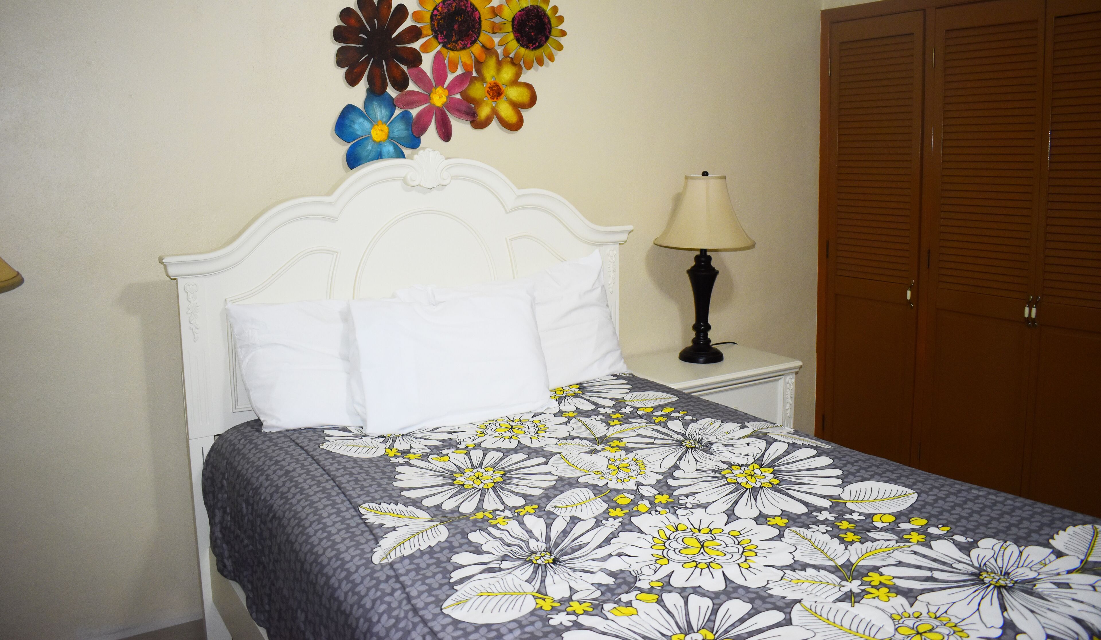 A neatly made bed with a floral bedspread, a wall-mounted flower arrangement, a bedside lamp, and a wooden wardrobe.