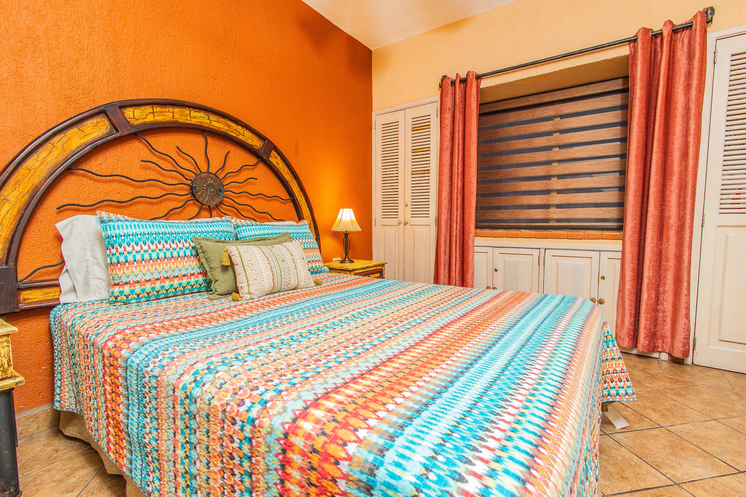 Une chambre avec un grand lit, une tête de lit ornée d’un motif solaire, une table de chevet avec une lampe et une fenêtre avec des volets.