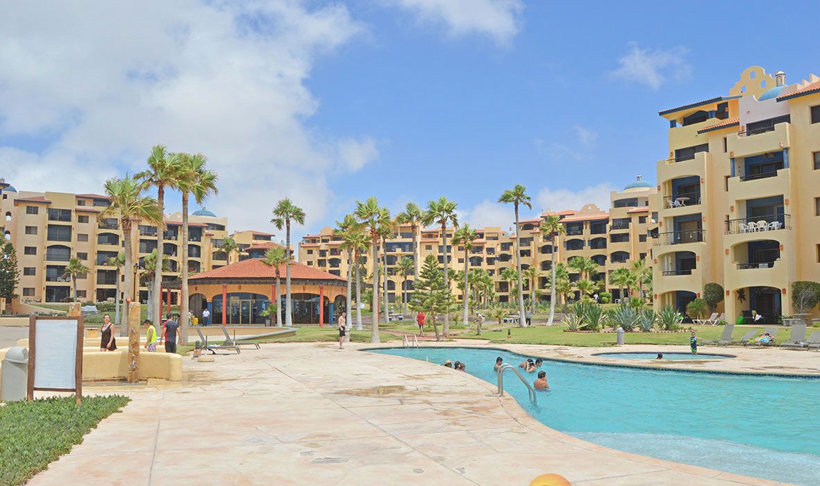A resort with multiple buildings, a central pool, palm trees, and a clear sky.