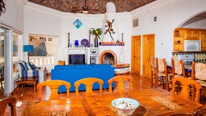 Een ruime woonkamer met een blauwe bank, een houten eettafel en stoelen, een open haard en een keuken met houten kastjes.