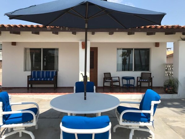An outdoor patio with a round table and chairs, a large umbrella, and a building with windows and a door.