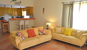 Ein Wohnzimmer mit einer Couch, zwei Sesseln, einem kleinen Tisch und einer Lampe. Es gibt eine Küchenzeile mit Holzausbau und einem Spülbecken.