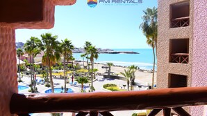 Blick vom Balkon auf einen Strandort mit Palmen, einem Pool und strahlend blauem Himmel.
