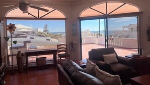 Ein Zimmer mit einer Couch, einem Esstisch, Stühlen, einem Deckenventilator und großen Fenstern mit Blick auf das Meer und Gebäude.