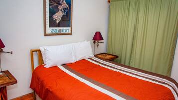 Un lit recouvert d’une couvre-lit rouge et blanche, un oreiller blanc, un tableau encadré représentant Puerto del Rosario, au Mexique, un rideau vert et une petite table avec une lampe.