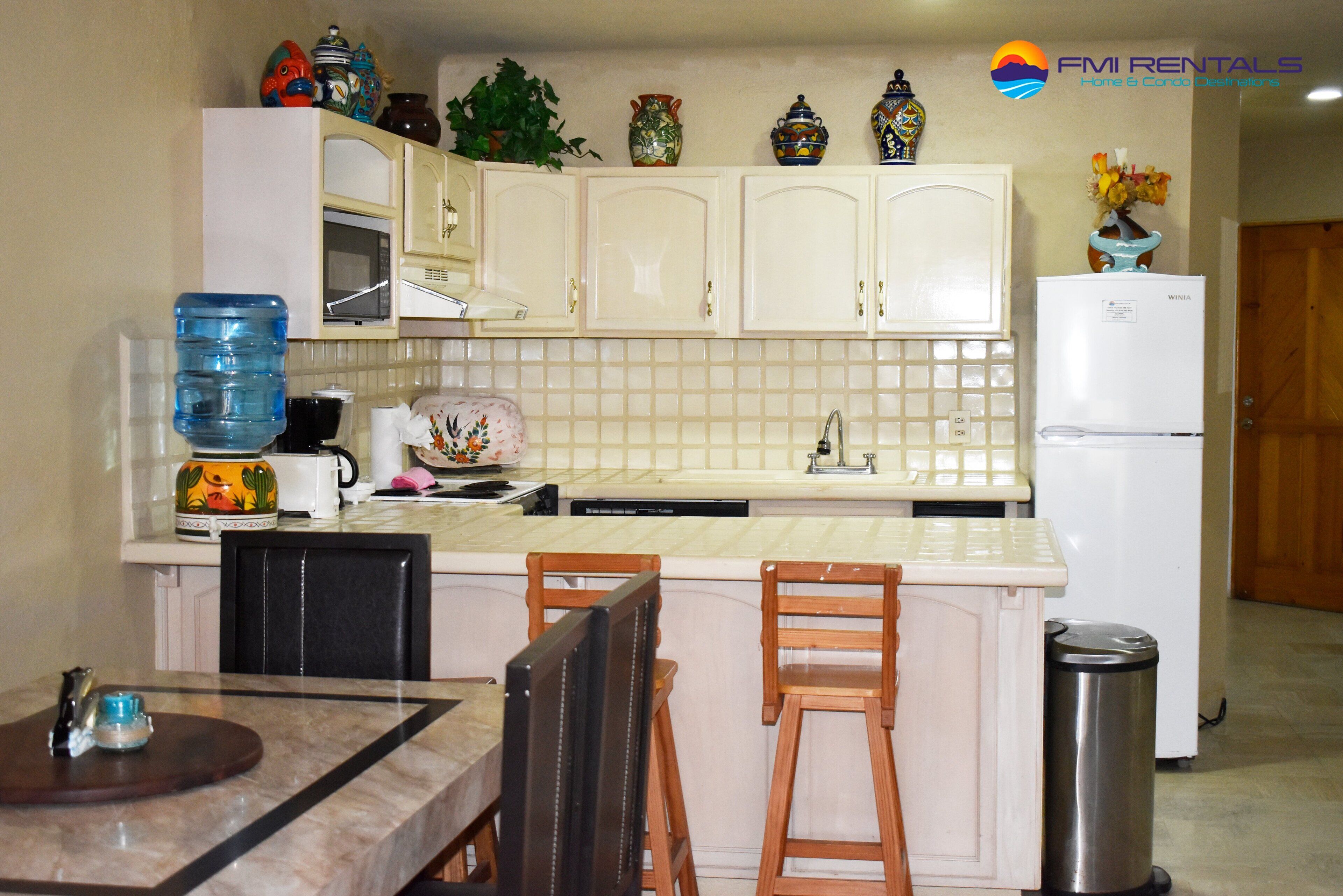 A kitchen with white cabinets, a microwave, a water dispenser, a sink, and a refrigerator. There is a dining table with chairs and a trash can.