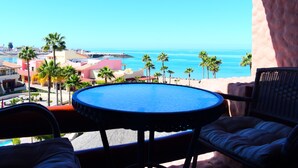 Een balkon met een ronde tafel en twee stoelen, met uitzicht op een strand met palmbomen en een helderblauwe lucht.