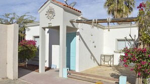 Un bâtiment blanc avec une porte bleue, un petit patio et des plantes à fleurs roses.