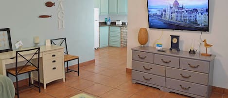 A living room with a television displaying a cityscape, a dining area with a table and chairs, and a kitchen in the background.