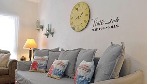 Un salon avec un canapé gris, des coussins décoratifs, une horloge murale et une citation inspirante.