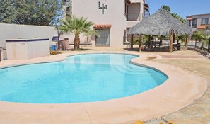 Une piscine à bord incurvé, entourée d’une terrasse en pierre. On y trouve également une structure à toit de chaume et un palmier à proximité.