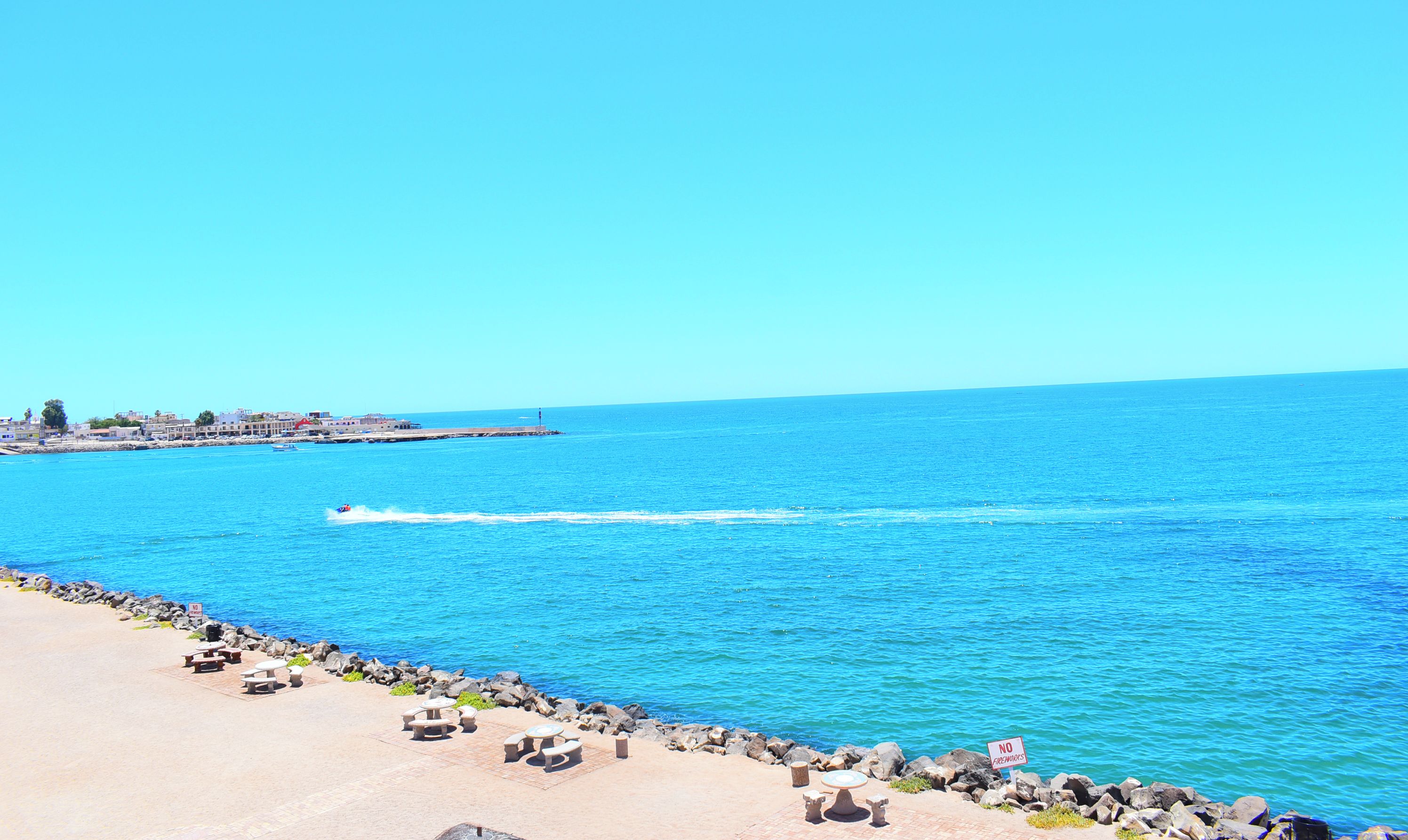 Una playa con cielo azul despejado, costa rocosa y un cartel que indica prohibido bañarse.