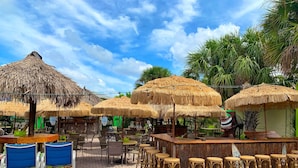 A beachside restaurant with thatched umbrellas, wicker bar stools, and outdoor seating.
