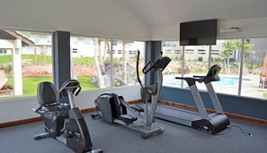 A gym with cardio machines and a TV screen, large windows with a view of a pool and greenery.