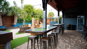Eine Terrasse im Freien mit einem langen Tisch und Barhocker, umgeben von Grünpflanzen und einem Pflasterboden.