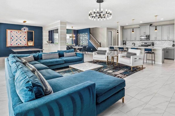 Una sala moderna con un sofá seccional azul, un comedor y una cocina con armarios blancos y una encimera de mármol.