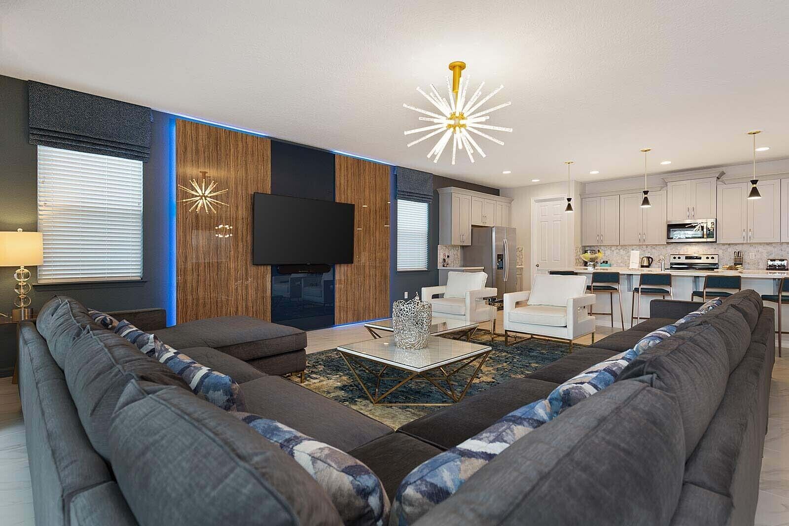 A modern living room with a sofa, a coffee table, and a TV. There is a kitchen area with white cabinets and a dining area with chairs.