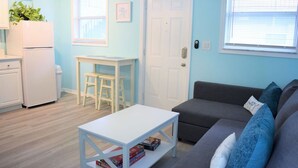 Una sala de estar con un sofá gris, una mesita de centro blanca y una pequeña mesa blanca con sillas. Hay una puerta blanca y una ventana con persianas.