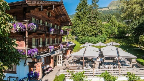 A traditional wooden chalet with balconies adorned with flowers, a terrace with outdoor seating, and a scenic view of a forested valley. - Landhotel Vordergrub (Kitzbuhel)