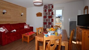 Un salon chaleureux avec un canapé rouge, une table à manger en bois et des chaises. Un téléviseur est posé sur un meuble en bois, et une fenêtre offre une vue sur les bâtiments environnants.
