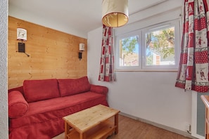 A room with a wooden wall, a red sofa, a wooden table, and a window with patterned curtains. - Studio abordable à 150m des remontées | Piscine (Les Deux Alpes)