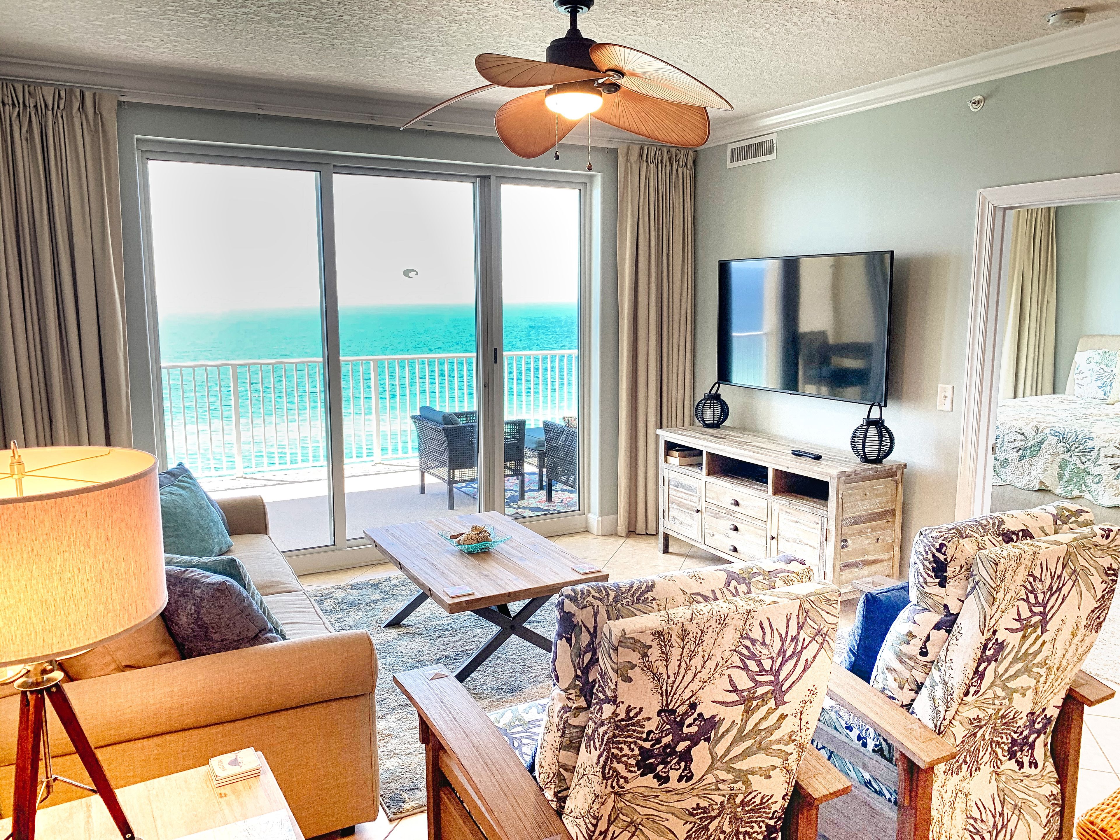A hotel room with a large sliding glass door leading to a balcony with a view of the ocean. There is a sofa, a coffee table, and two chairs with floral upholstery. A flat-screen TV is mounted on the wall, and a ceiling fan is visible.