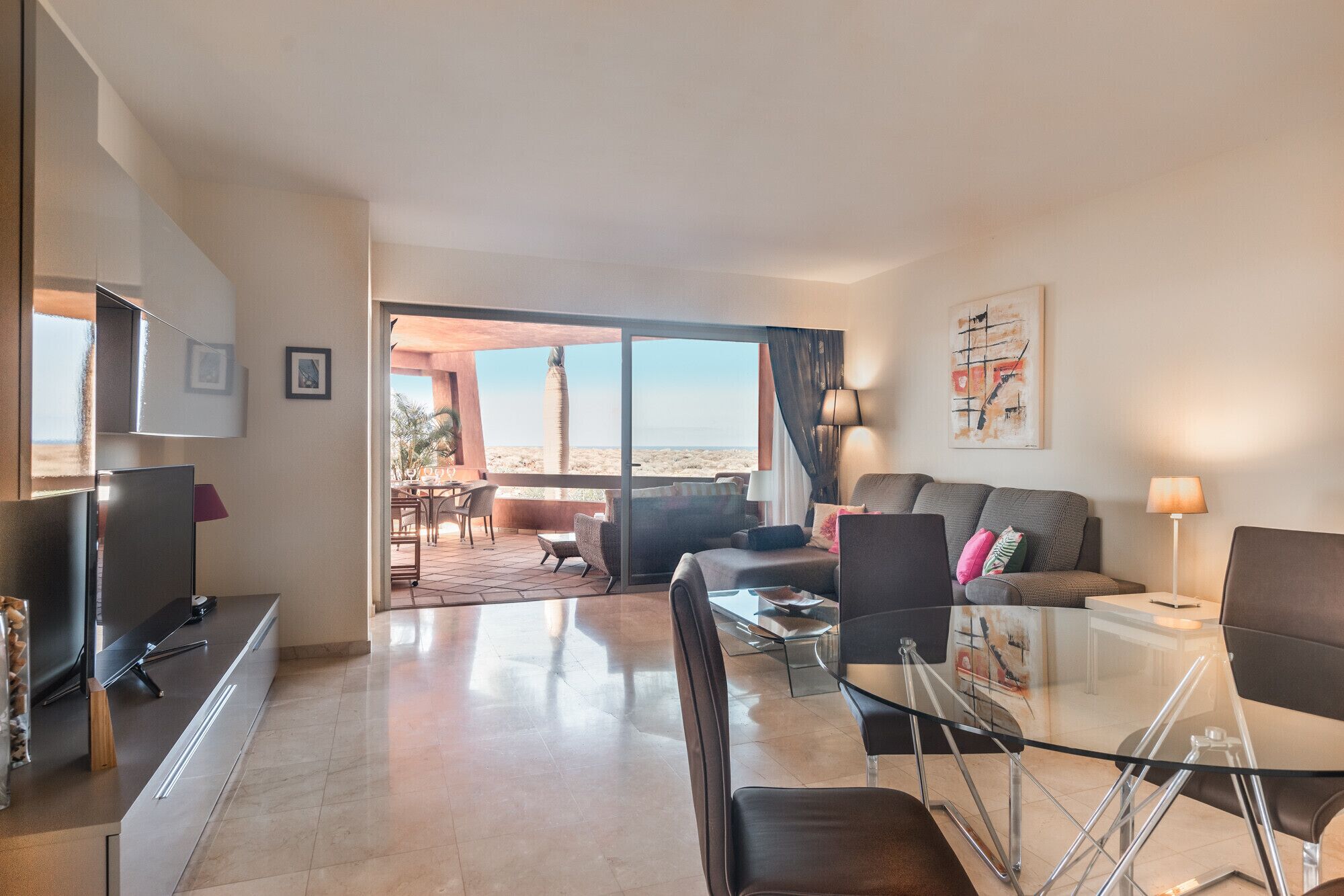 Een moderne woonkamer met een glazen eettafel, een bank en een televisie. Er is een balkon met uitzicht op het strand.