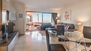 A modern living room with a glass dining table, a sofa, and a TV. There is a balcony with a view of the beach. - Bahia - Beneharo 1.1 DELUXE SEA VIEW 2B (Arona)