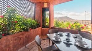 A terrace with a table set for dining, surrounded by lush plants and a view of a desert landscape. - Bahia - Beneharo 1.1 DELUXE SEA VIEW 2B (Arona)