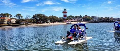 A watercraft with two people on it, a lighthouse, and a beach with buildings in the background.