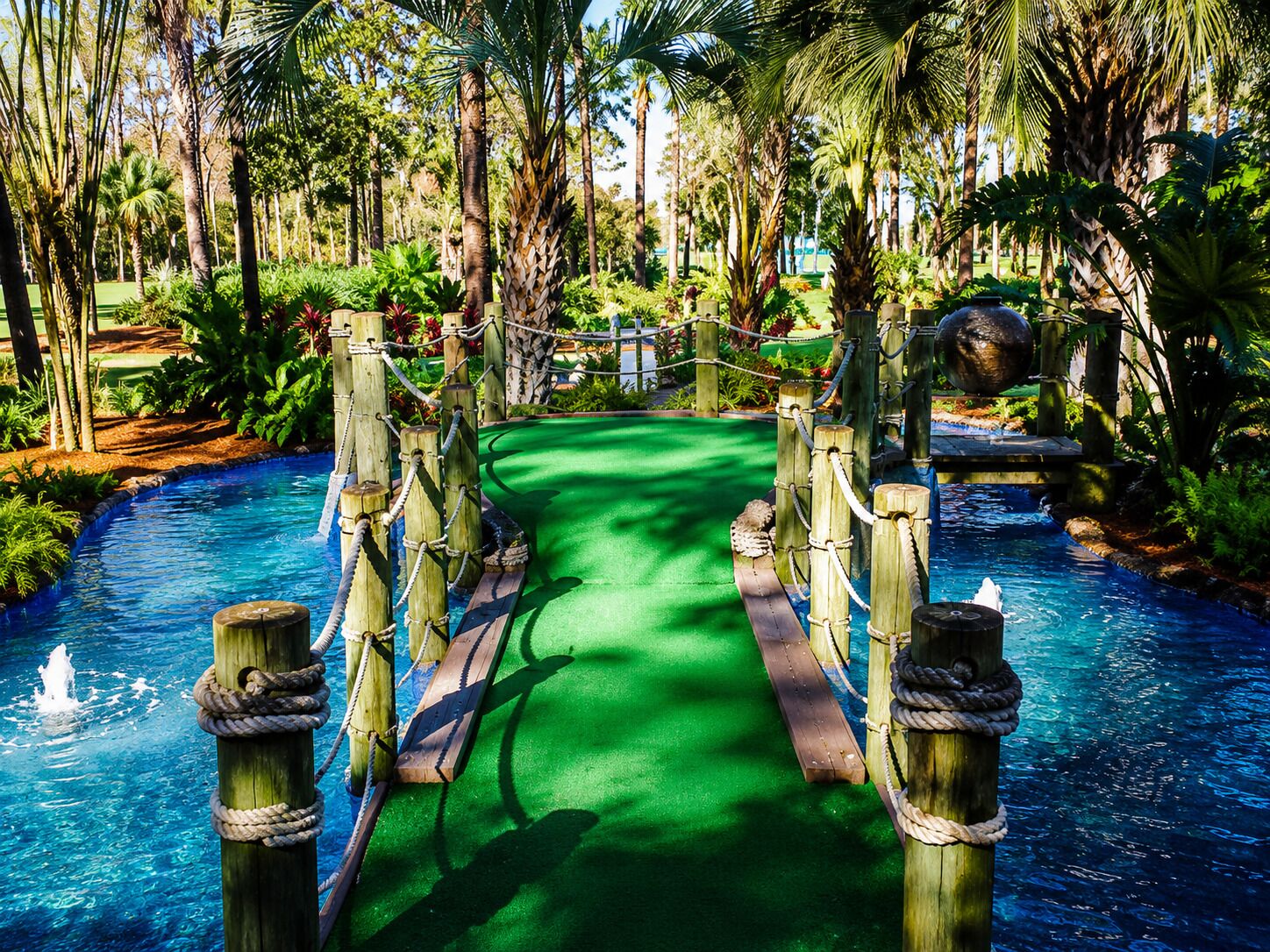 A wooden bridge over a water channel with a green carpet path, surrounded by lush tropical vegetation.