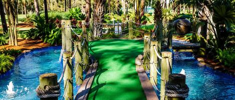 A wooden bridge over a water channel with a green carpet path, surrounded by lush tropical vegetation.