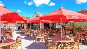 Un espace extérieur aménagé avec des tables et des chaises en bois, des parasols rouges et un ciel d’un bleu éclatant.