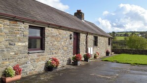 A stone building with a sloped roof, a chimney, and a door with a window above it. There are flower pots with flowers on the ground in front of the building. - 3 bedroom accommodation in Pontyates, near Kidwelly (Pontyates, near Kidwelly)