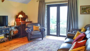 A living room with a leather sofa, a dark armchair, a television, a wooden cabinet, and a large window with curtains. - 3 bedroom accommodation in Pontyates, near Kidwelly (Pontyates, near Kidwelly)