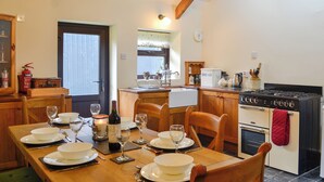 A dining area with a wooden table set for a meal, a kitchen with a stove and oven, and a window with blinds. - 3 bedroom accommodation in Pontyates, near Kidwelly (Pontyates, near Kidwelly)