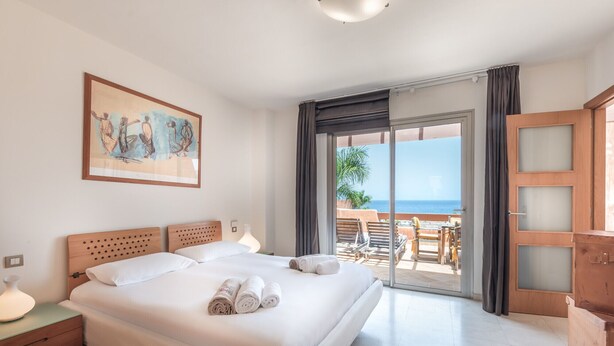 Ein Hotelzimmer mit Bett, einem Gemälde, einem Nachttisch, einer Lampe und Blick auf das Meer.