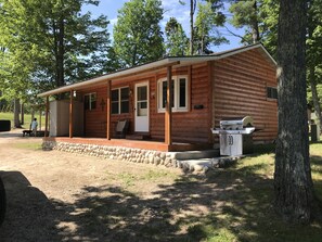A wooden cabin with a porch, a grill, and a stone wall. - Modern 2 Bedroom 1 Bath Just Steps From The Au Train River (Au Train)