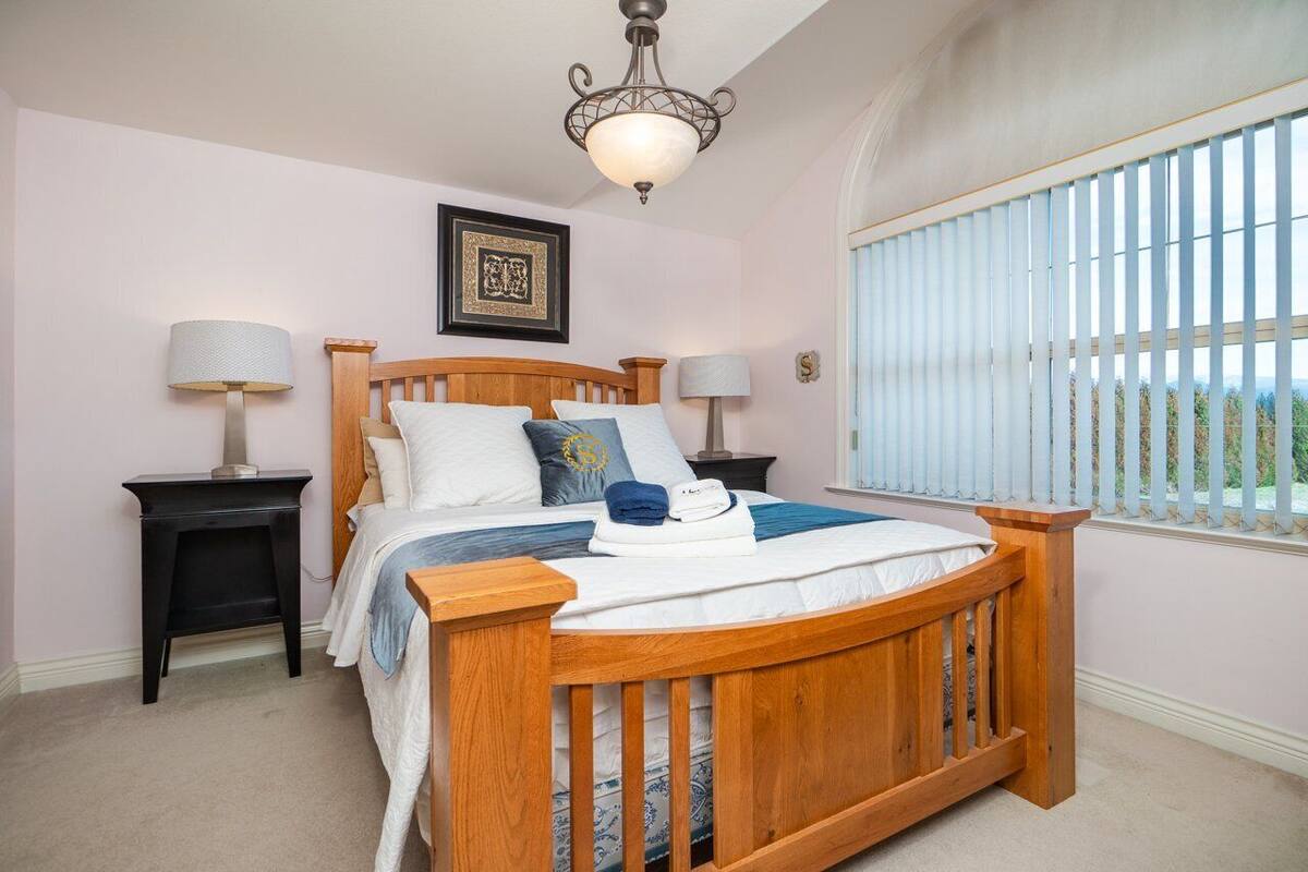 A bedroom with a wooden bed, two bedside tables, a ceiling light fixture, and a window with blinds.