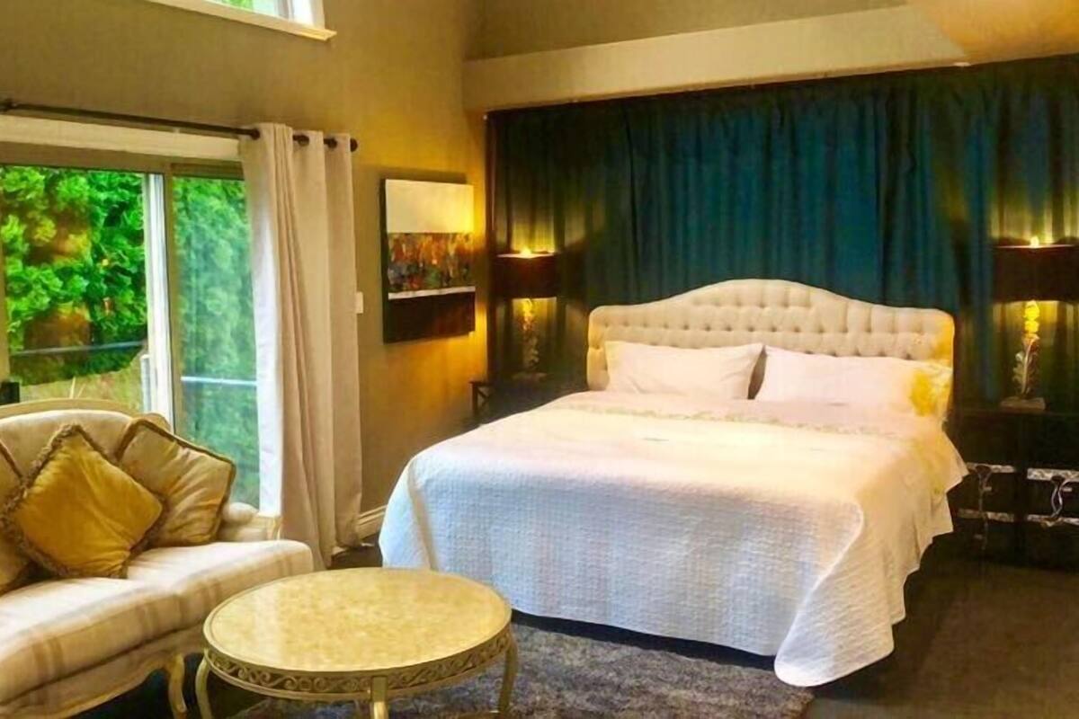 A bedroom with a large bed, a sofa, a small round table, and a view of greenery outside.