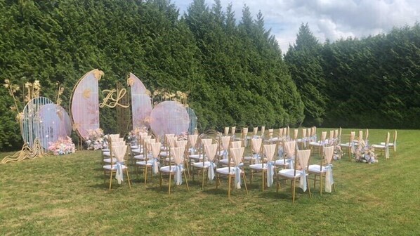 An outdoor wedding setup with rows of chairs and tables, decorated with floral arrangements and a large, ornate arch.