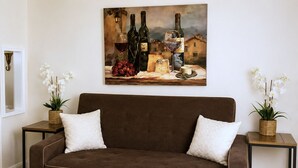A brown sofa with two white cushions, flanked by two small tables with wicker baskets and white orchids. A painting of wine bottles and grapes hangs above. - Patio + BBQ + Downtown Access | Paso Robles | Vine Street Guest House (Paso Robles)