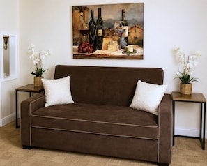 Ein braunes Sofa mit zwei weißen Kissen, das von zwei kleinen Tischen mit Korbwannen und weißen Orchideen flankiert wird. Darüber hängt ein Gemälde mit Weinflaschen und Trauben.